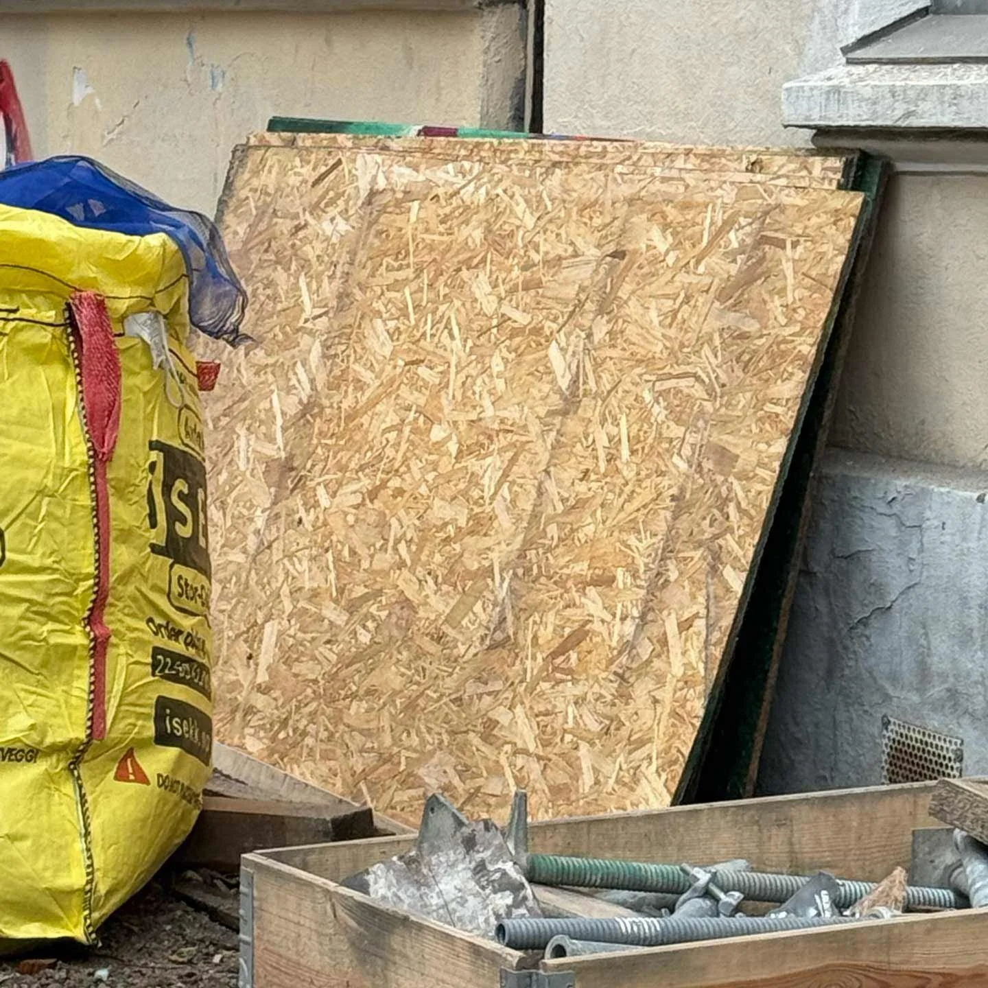 Construction materials including a large sheet of oriented strand board, a bag of fast-building mix, and a wooden box with metal bolts and screws, near a wall with concrete and siding textures.