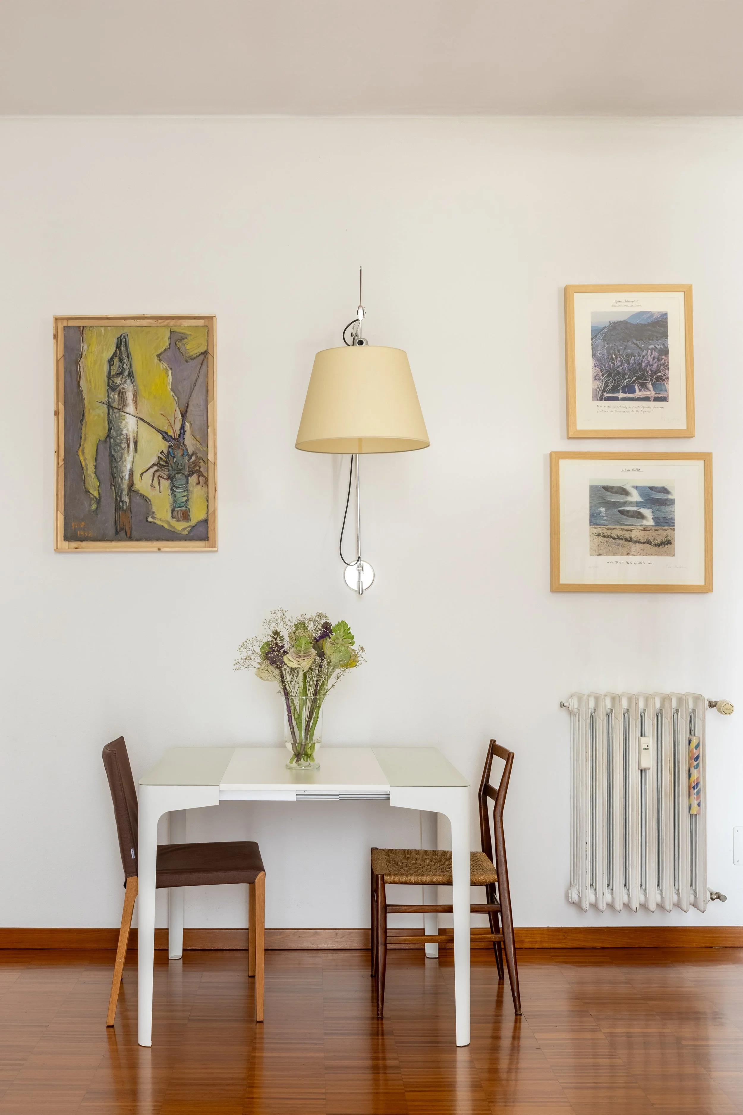 Interno di una stanza con pareti bianche, tavolo bianco con vaso di fiori, due sedie di diverso stile, quadro con disegni di paesaggi e quadro con un dipinto astratto, lampada appesa, termosifone.