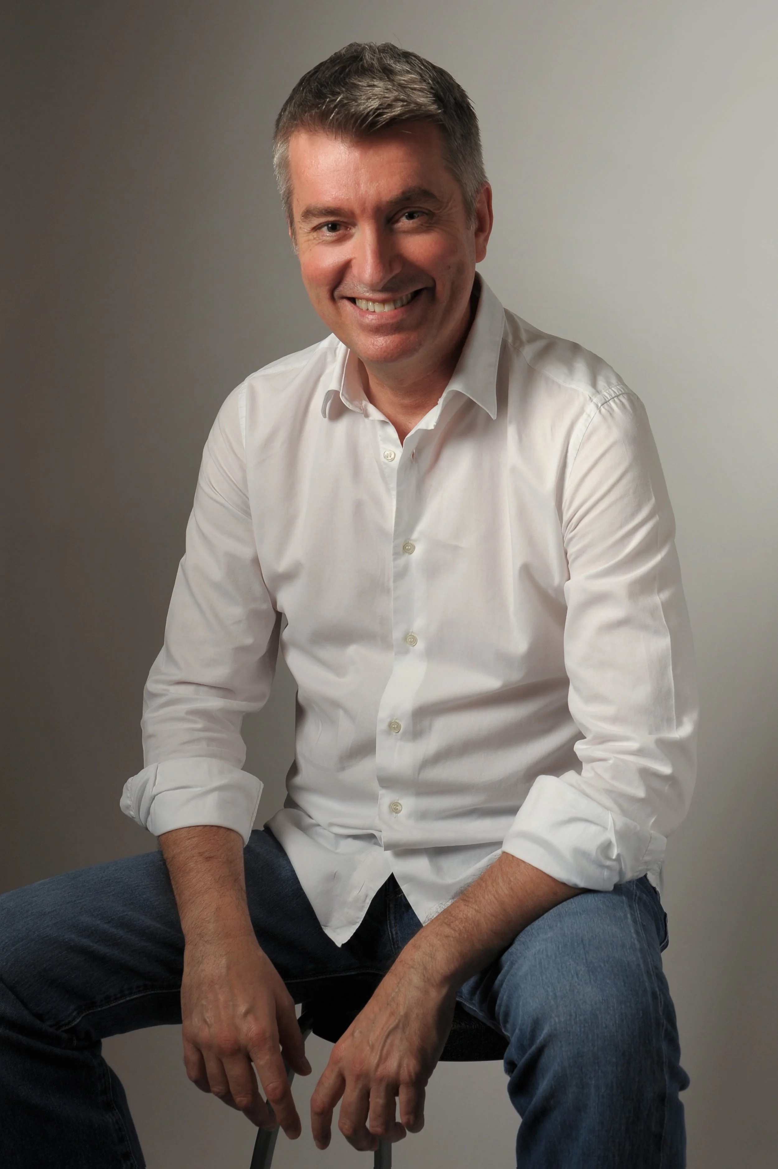 Uomo sorridente seduto con camicia bianca e jeans, sfondo neutro.