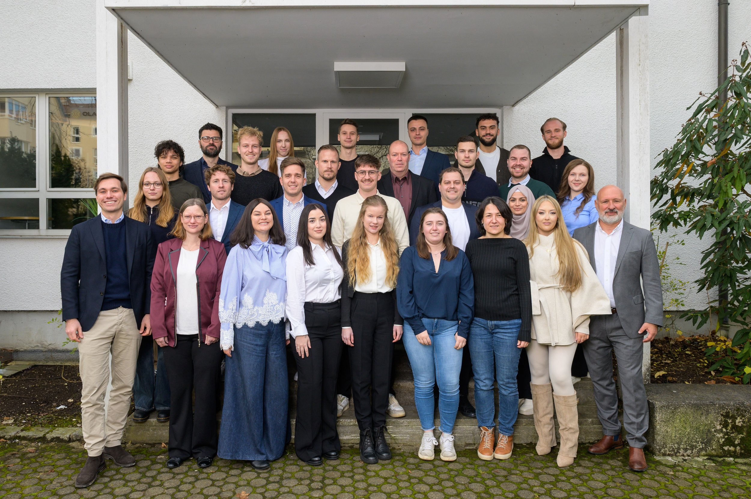Gruppenfoto von Menschen vor einem modernen Gebäude mit weißer Fassade, inklusive Männer und Frauen unterschiedlichen Alters und ethnischer Hintergründe, die sich für ein Gruppenfoto versammelt haben.