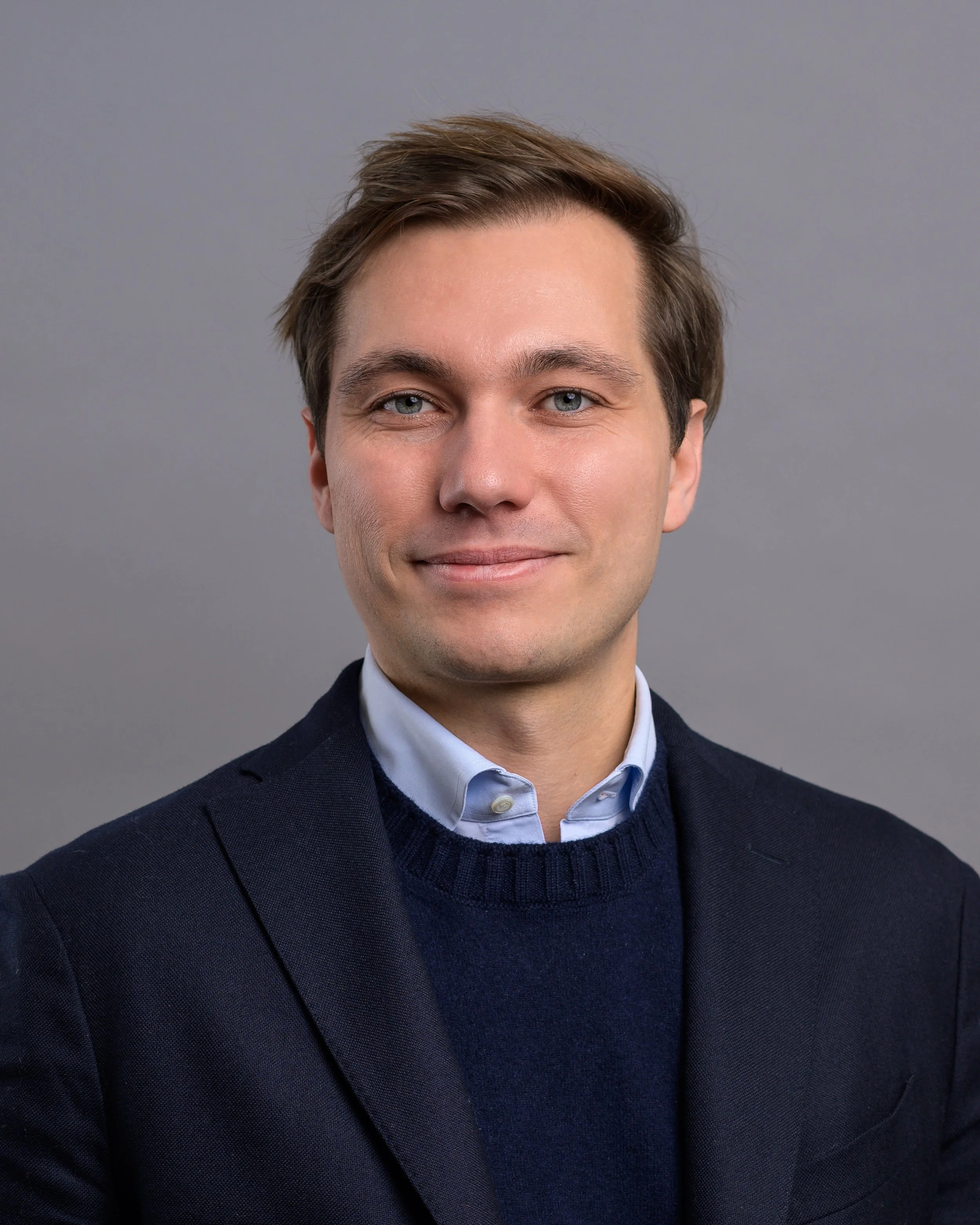 Porträt eines Mannes mit braunen Haaren, blauen Augen, dunkelblauer Jacke und weißem Hemd gegen grauen Hintergrund.