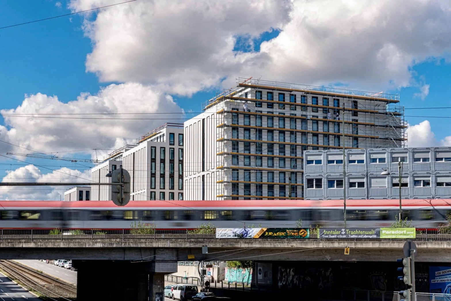 Moderne, weiße Bürogebäude im Bau, ein Zug fährt vorbeigefahren, unter einer Brücke mit einigen Autos. Der Himmel ist teilweise bewölkt.
