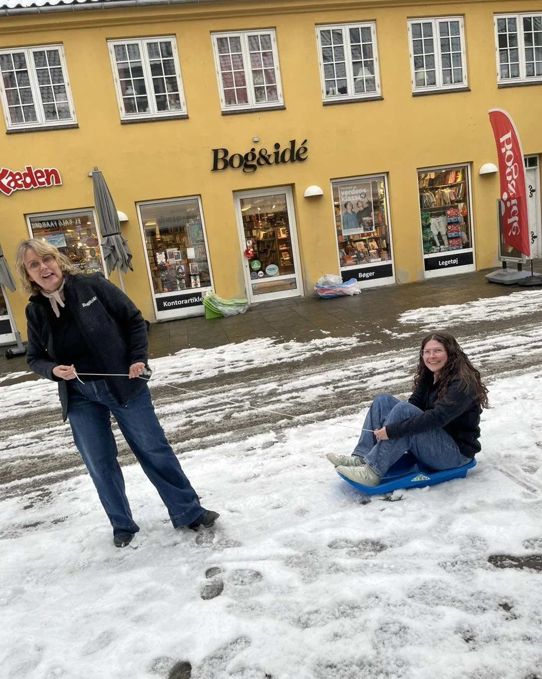 Two women in winter clothing are outside on snowy and icy ground in front of a yellow building with bookstore windows. One woman, seated on a plastic sled, is smiling, while the other woman, standing, is holding the sled's rope.