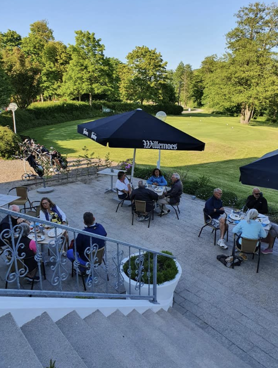 People dining outdoors on a patio overlooking a golf course with green fairways and trees, shaded by large umbrellas.