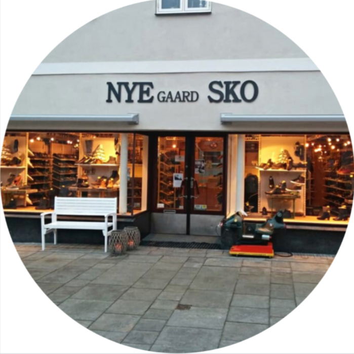 Storefront with sign 'NYE GAARD SKO' selling shoes, displayed in windows with warm lighting, featuring a white bench and outdoor equipment in front.