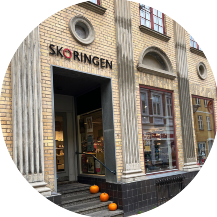 Exterior of a brick building with the sign 'SKORINGEN' above the entrance, featuring tall windows with red frames, decorative circular accents, and four pumpkins on the steps leading into the store.