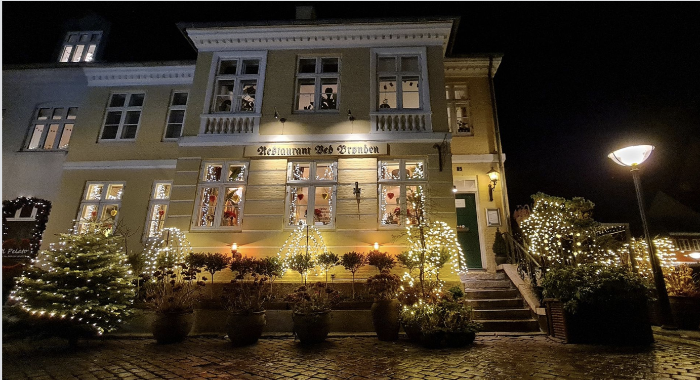 The exterior of a yellow, two-story building decorated with Christmas lights and ornaments. The building has a sign that reads 'Restaurant Ned Bronden' and is adorned with potted plants and trees with string lights. There is a staircase leading to a green door on the right, and a streetlamp on the right side illuminating the scene at night.