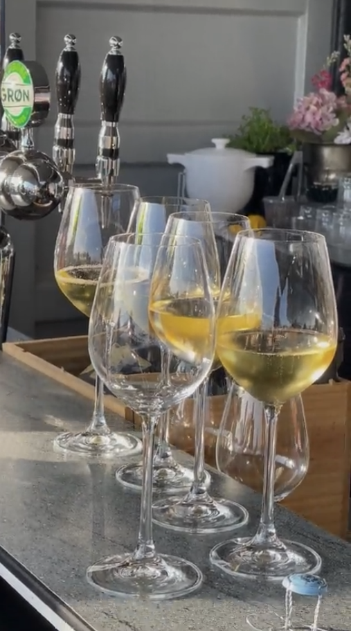 Four wine glasses filled with white wine on a bar counter, with a beer tap in the background and a kitchen setting.
