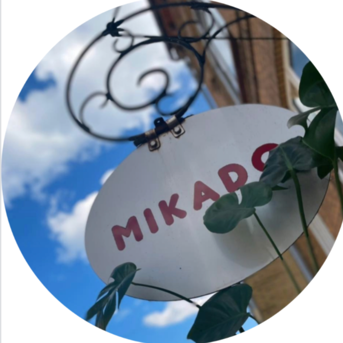 White oval sign with red text reading "MIKADOS" hanging outdoors against a blue sky with clouds, surrounded by green leaves.