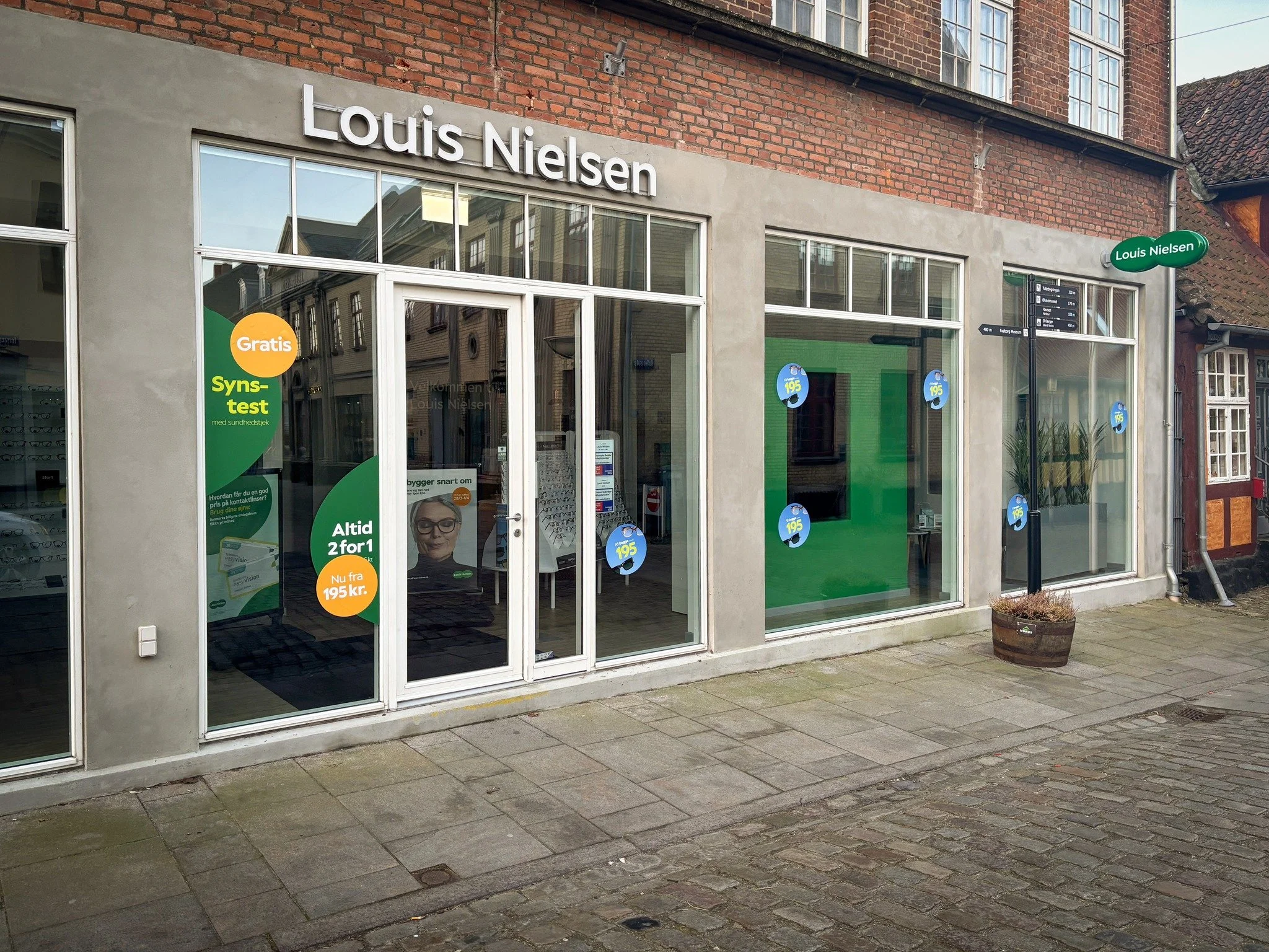 Facade of a Louis Nielsen store with large glass windows and door, signs advertising free eye tests and special offers, in a building with a brick upper floor and red-tiled roof, situated on a cobblestone sidewalk.