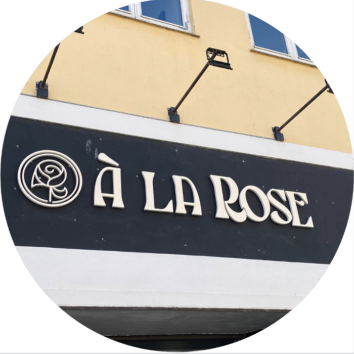 Storefront sign for 'À La Rose' with a simple rose logo on a black background, located under yellow building with windows.