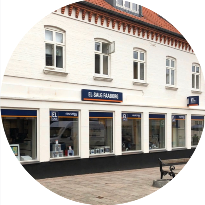 Outside view of a white building with a sale sign reading 'EL-SALG FAABORG' and multiple windows, with the store named 'Euronic' in the display windows and a bench on the sidewalk.