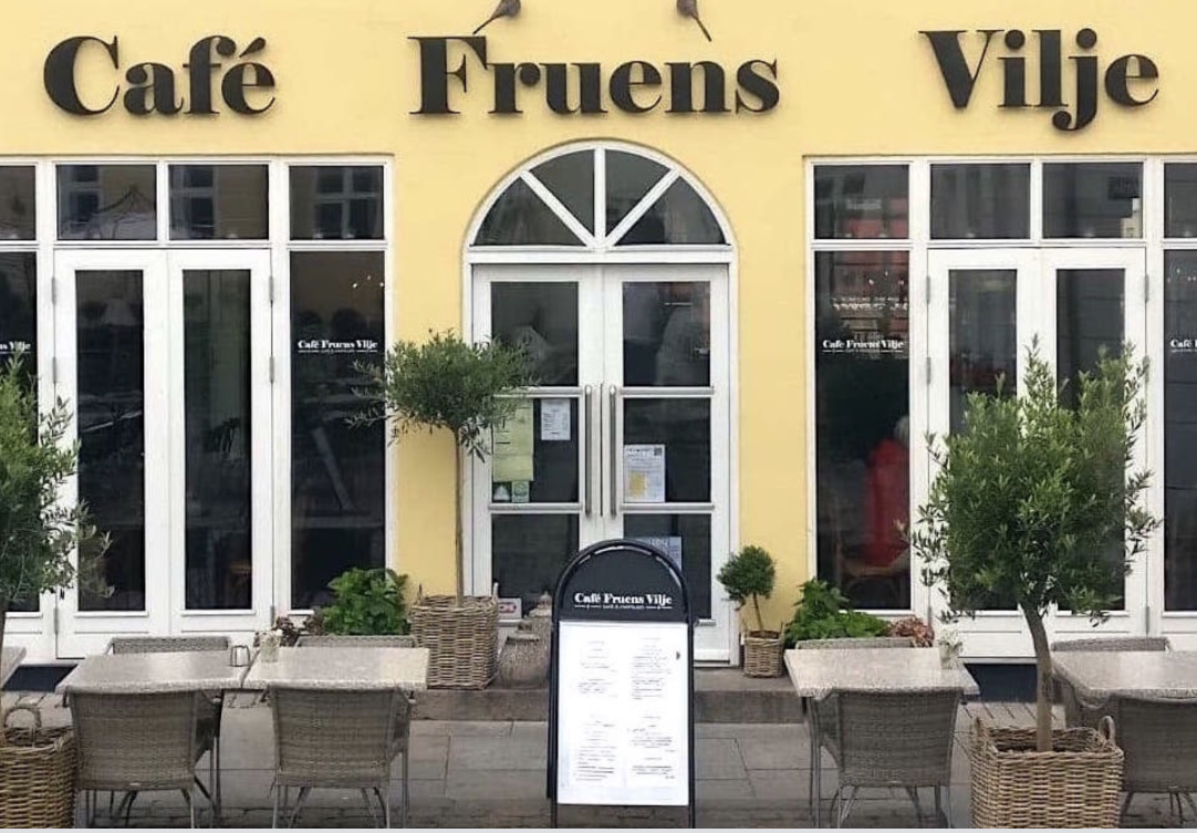 Exterior of Café Fruens Vilje with yellow facade, large windows, potted trees, and outdoor seating with wicker chairs and tables.