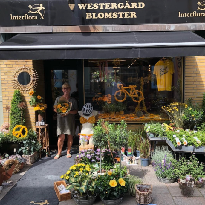 A woman stands outside a flower shop named Westergård Blomster, holding a bouquet of yellow flowers. The shop window displays a bicycle arrangement and a yellow t-shirt. The front area has various plants and flowers, with some sunflowers and decorative items like a sunflower wreath and a small pep rally sign.