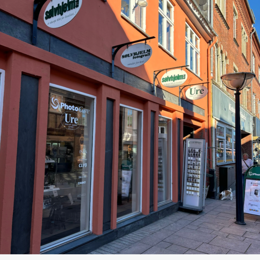Street view of a photography store with large glass windows, multiple signs that say 'Sølvhjelm Fotografi' and 'Ure', and a street lamp. A person and a dog are seen near the storefront.