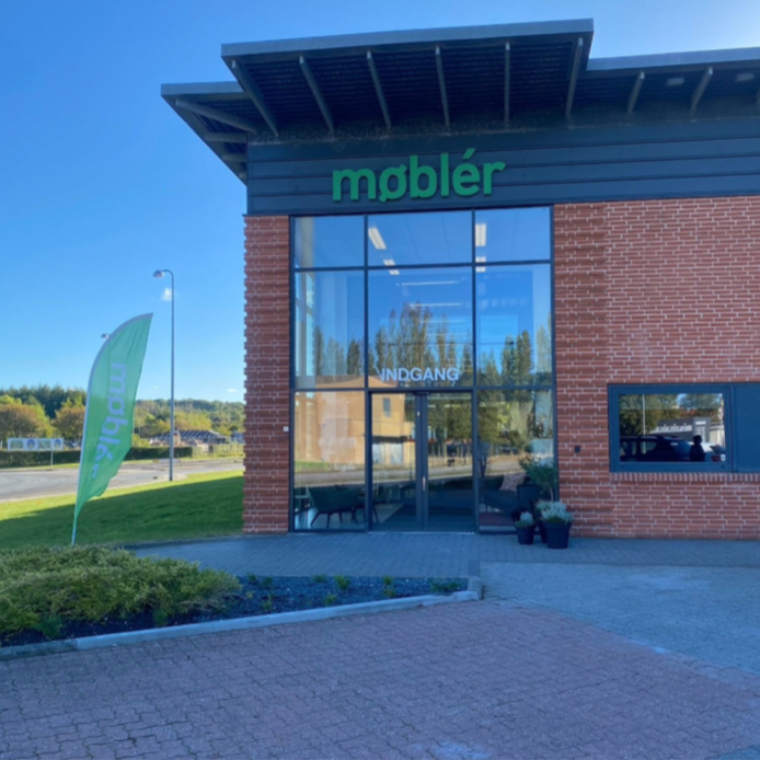 Exterior view of a store named Møbler with glass doors and windows, a green banner on the side, and a sign indicating the entrance.