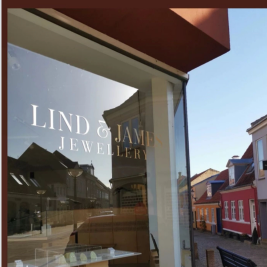 Storefront window with signs for Linda & James Jewellery, reflecting surrounding buildings and street outside.