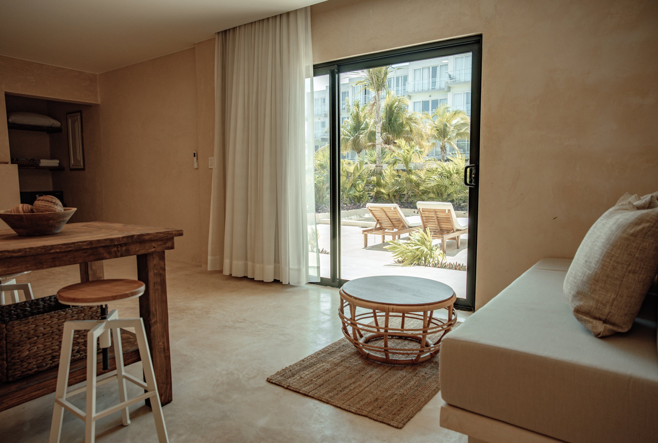 Living room with sliding glass door leading to outdoor patio with lounge chairs, palm trees, and neighboring buildings, beige walls, a wicker coffee table, a white sofa with pillows, a woven rug, and a wooden dining table with stools.