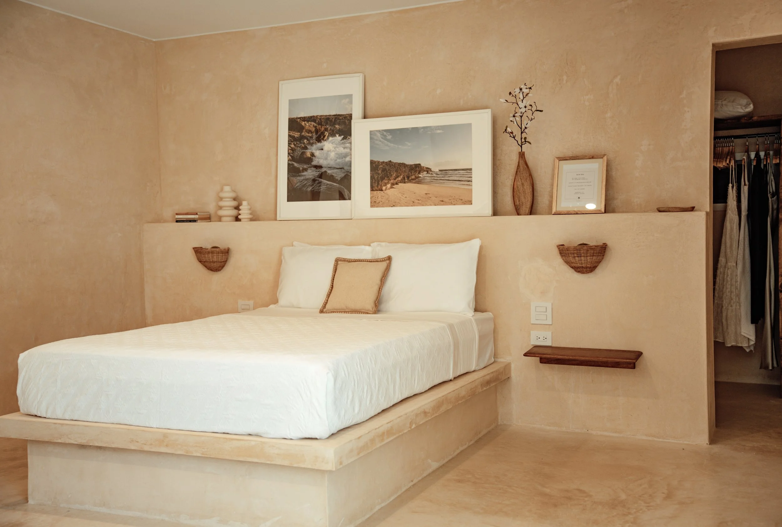 A minimalist bedroom with beige walls and flooring, featuring a bed with white bedding and a decorative pillow. Behind the bed, there are three framed landscape photographs, a tall vase with branches, and small decorative objects on a narrow ledge. T