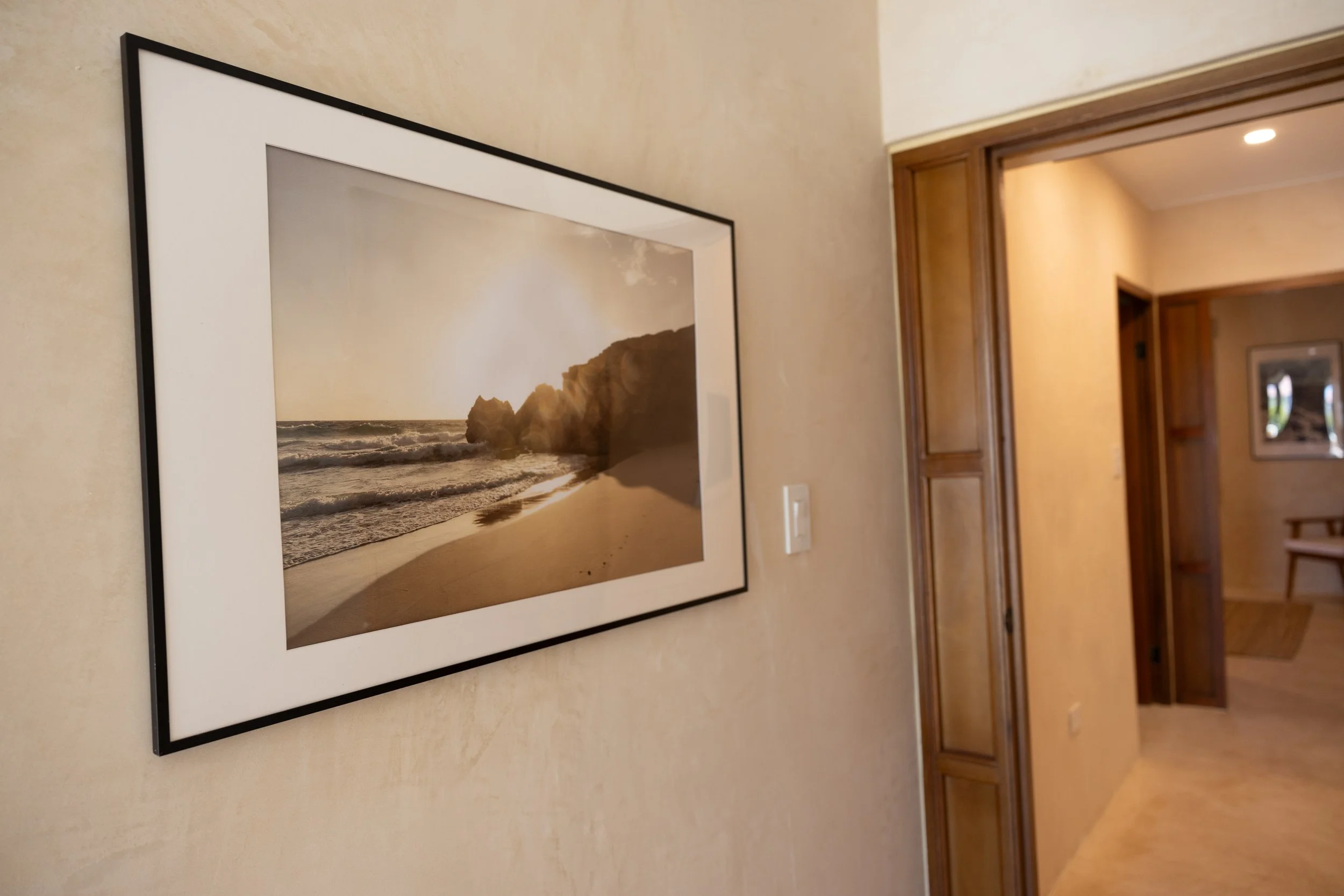 View of a framed beach photograph hanging on a beige wall in a hallway with wood trim and a door leading to another room.