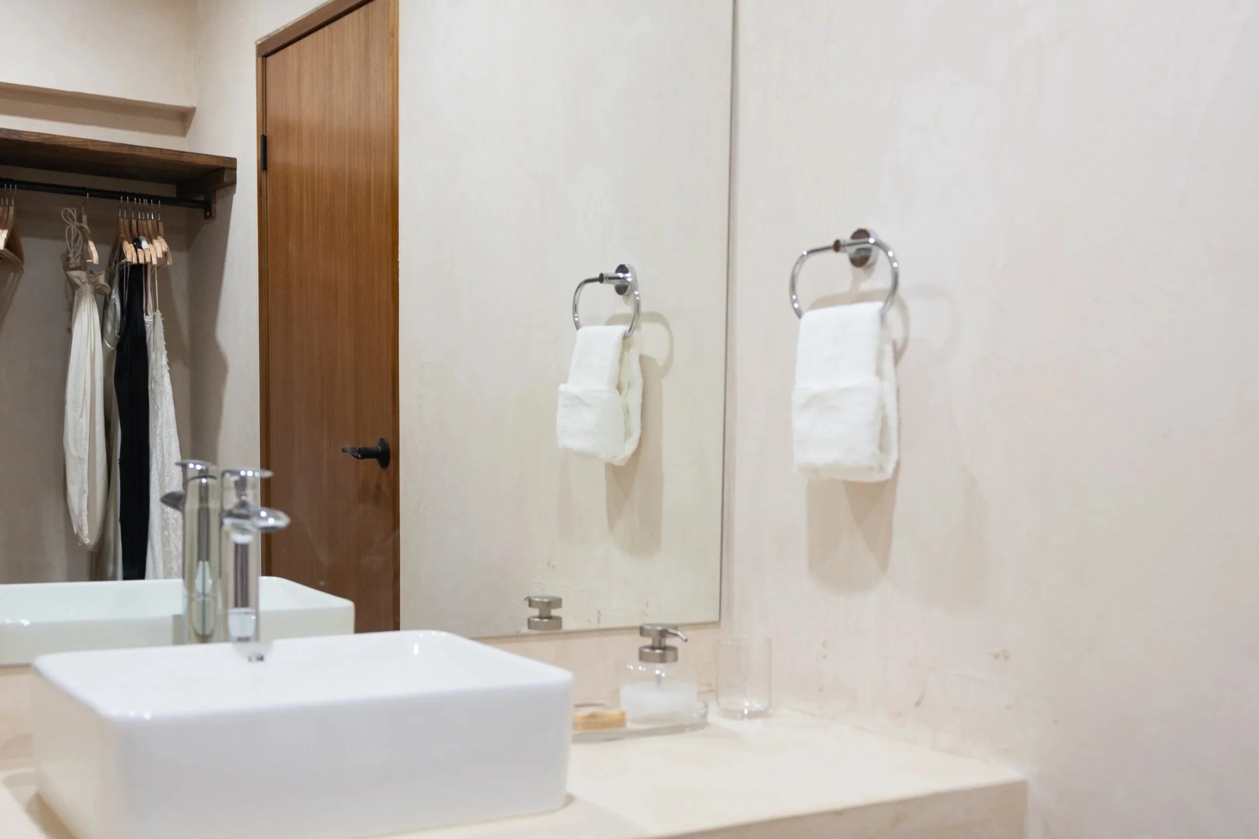 Bathroom with a white sink, a mirror, hand towels on chrome rings, a soap dispenser, and a wooden door.