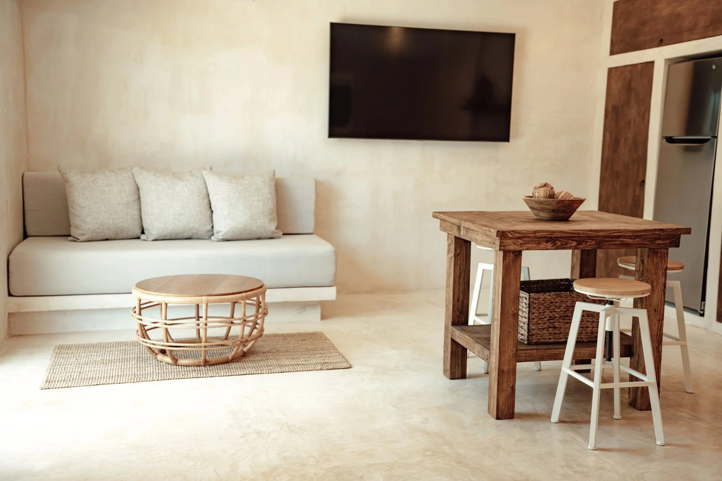Living room with a beige couch and three pillows, a rattan coffee table on a woven rug, a wall-mounted flat-screen TV, and a wooden dining table with two white barstools, a basket beneath the table, and a small bowl on top.