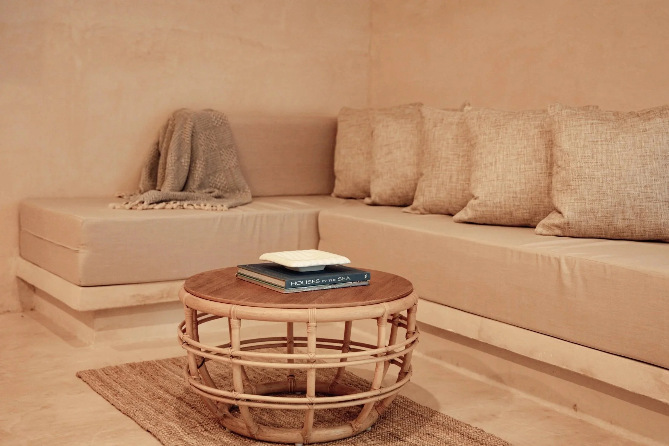 A beige living room corner with a sectional sofa, beige cushions, a woven throw blanket, and a round rattan coffee table with a book and a cushioned item on top, on a small rug.