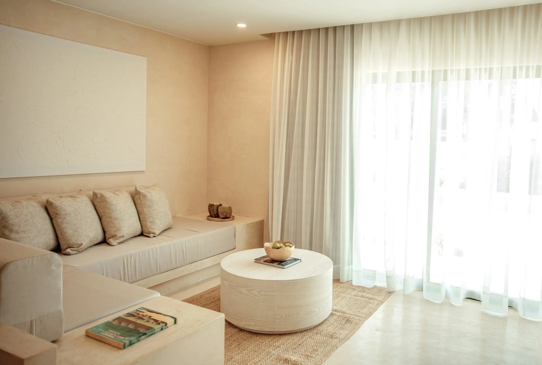 A cozy living room with beige walls, a cream sofa with pillows, a round white coffee table with a bowl and books, a large window with sheer white curtains, and soft lighting.