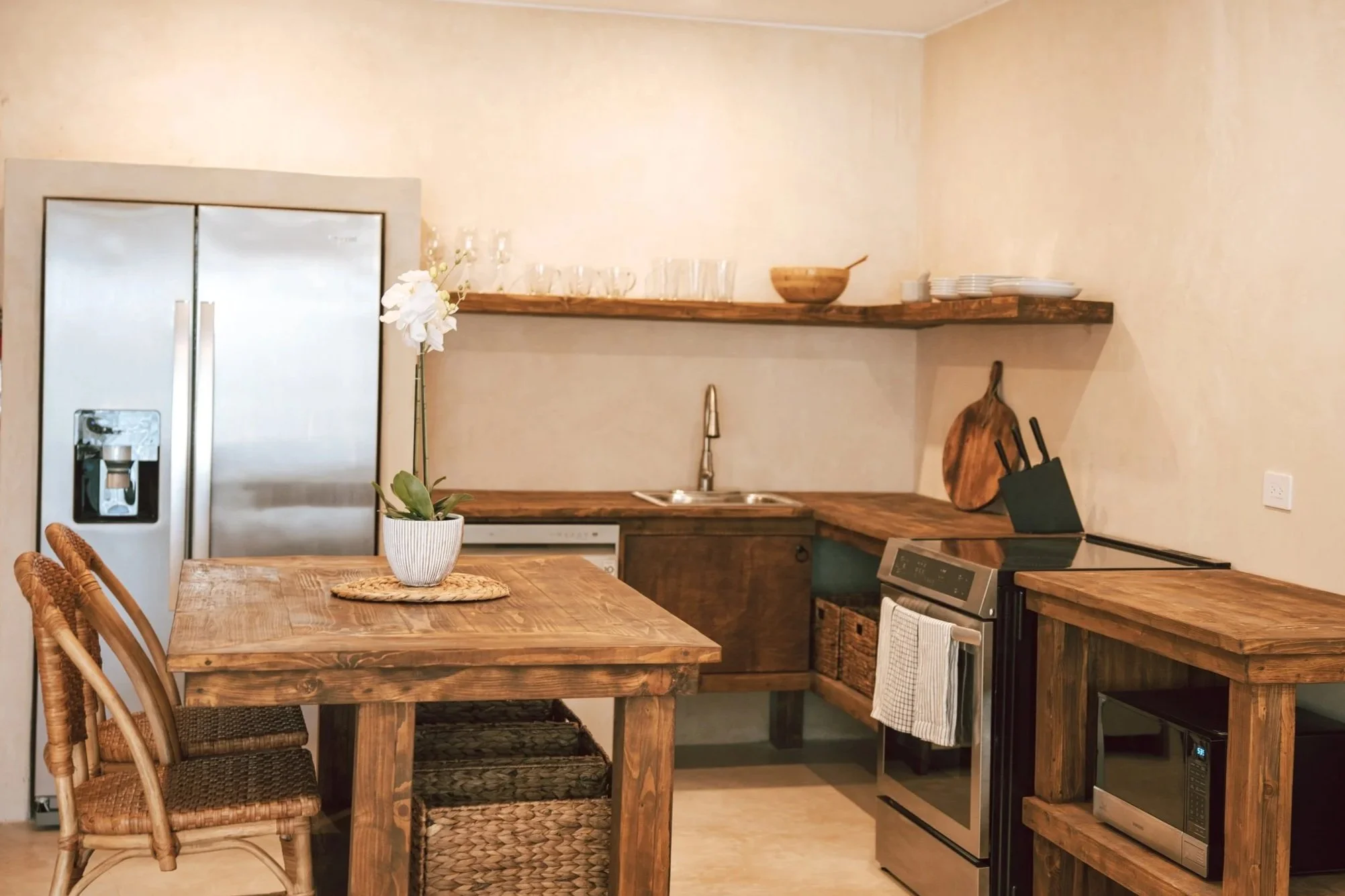 Kitchen with wooden dining table, chairs, a potted orchid, refrigerator, open shelves with glasses and bowls, microwave, oven, and cutting boards.