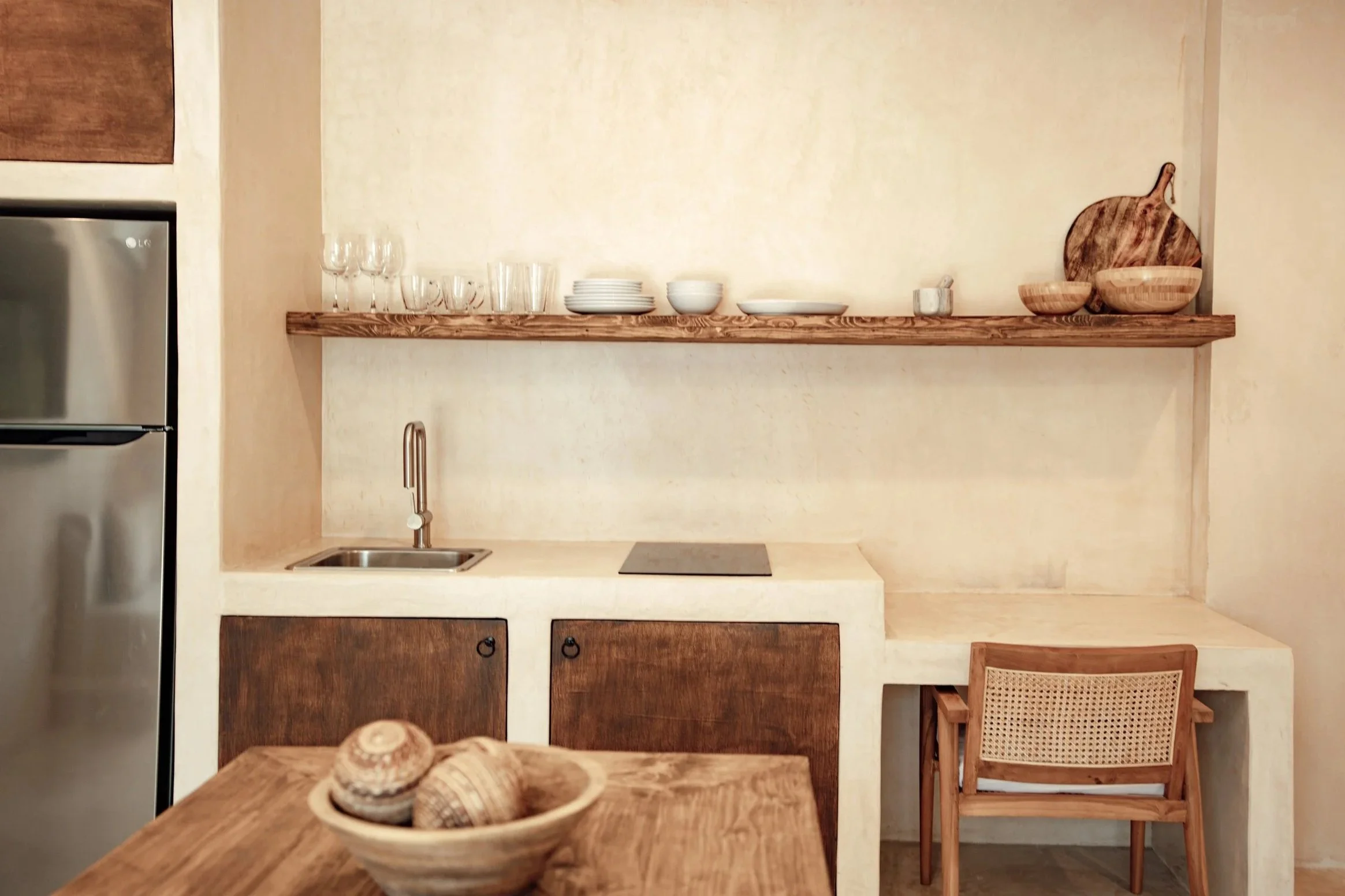 Minimalist kitchen with open wooden shelves, small sink, and a wooden chair at a built-in desk, decorated with bowls and glasses.