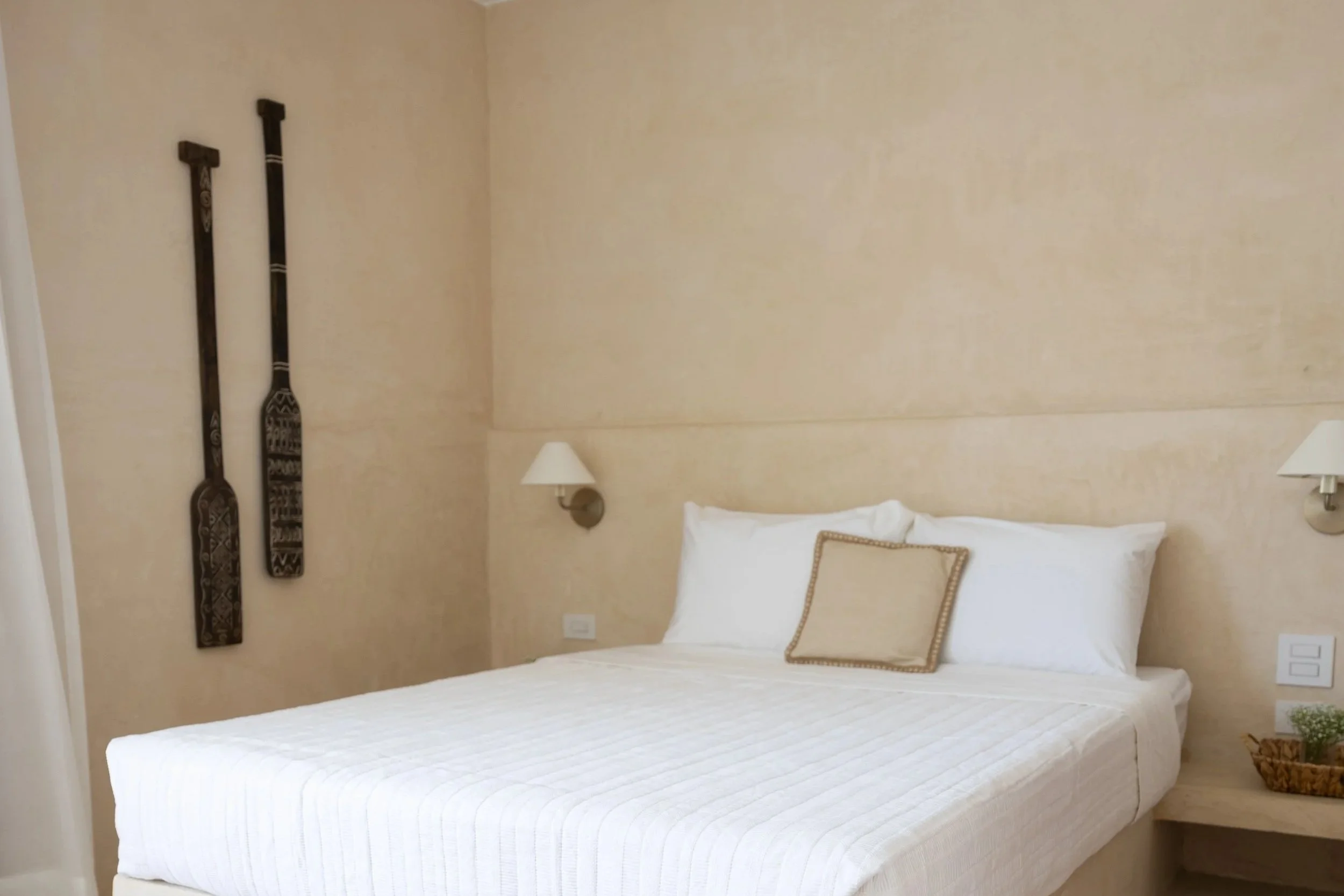 A neatly made bed with white linens and a beige decorative pillow, two wall-mounted reading lamps on either side, beige painted walls, and traditional African wooden paddles hanging on the left wall.