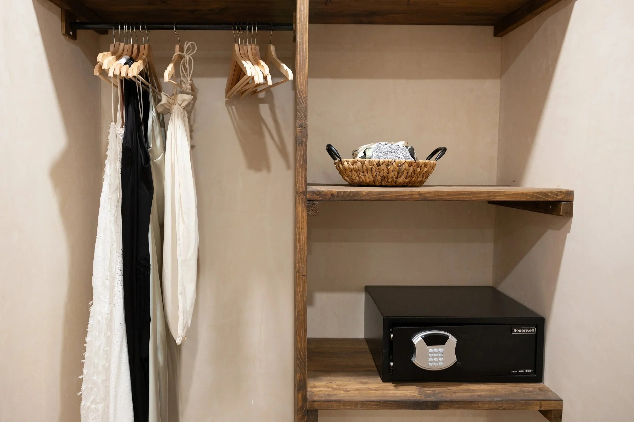 Open closet with hanging dresses, a woven basket on a shelf, and a black safe on the bottom shelf.