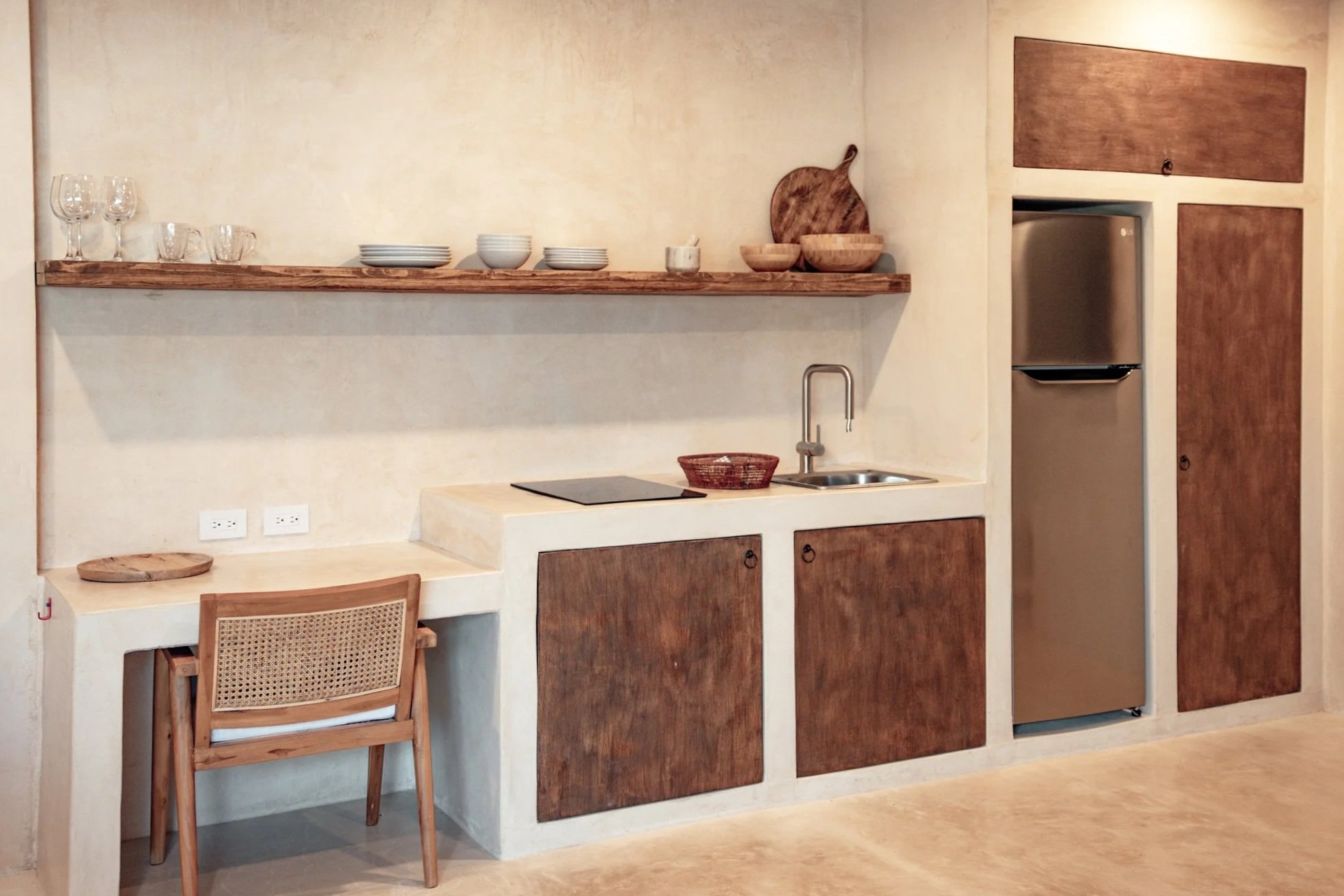 A minimalist kitchen with a wooden shelf holding glasses, bowls, and cutting boards. Below the shelf is a counter with a cooktop, a small bread basket, and a sink. The cabinetry and wall accents are made of wood, and there is a small wooden desk with