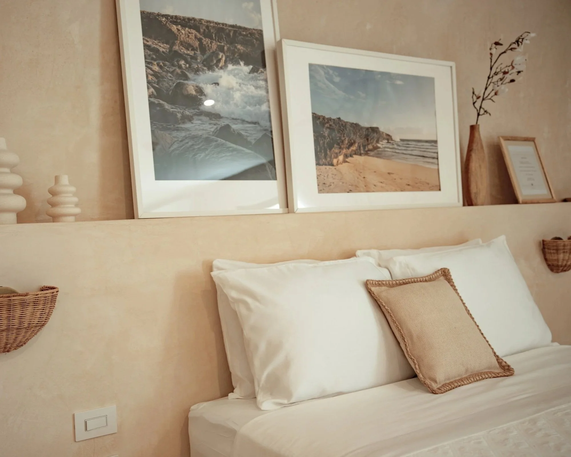 Bedroom with white bed, beige pillows, wall art photos of beach and ocean, wooden vase with branch, framed picture, and woven baskets on a beige wall.