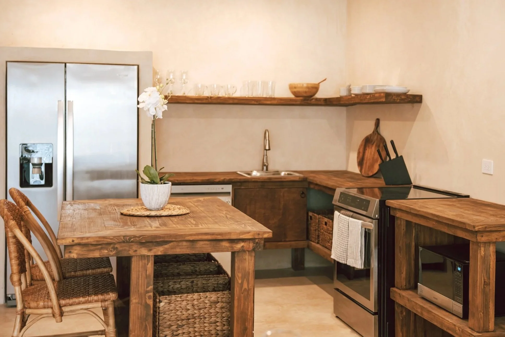Rustic kitchen with wooden countertops, a stainless steel refrigerator, a stove, a microwave, and a wooden dining table with wicker chairs. A pot with an orchid is on the table, and open shelves hold dishes and glasses.