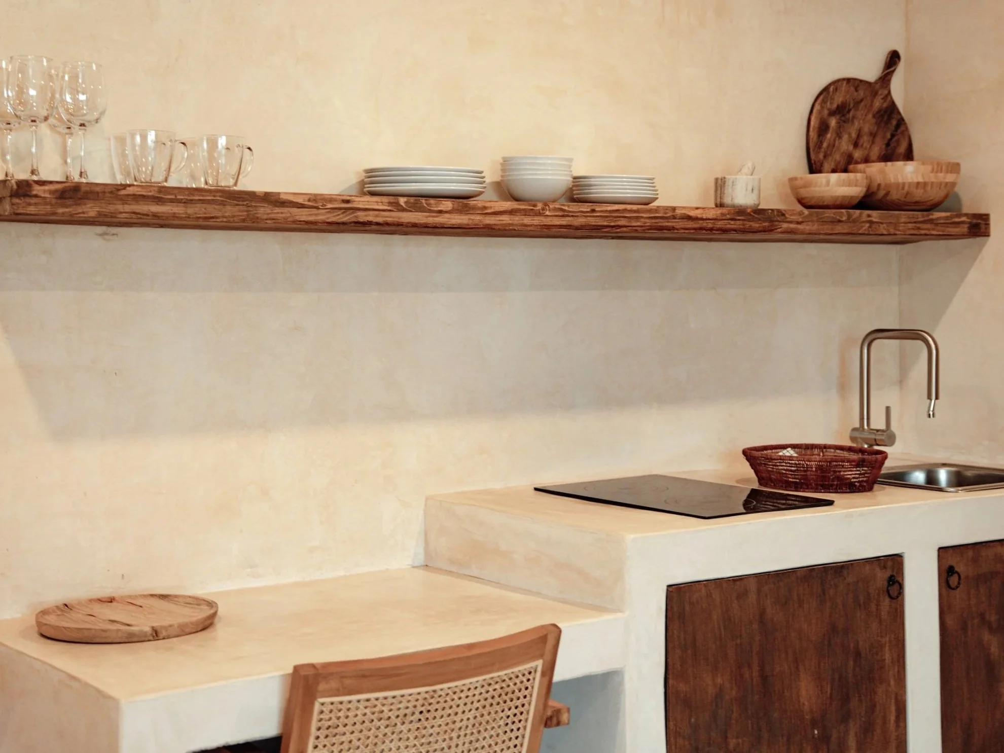 Minimalist kitchen with a wooden shelf holding glassware, plates, bowls, and cutting boards. Below is a countertop with a small sink, a wooden bowl, and a cooktop, with a wooden cabinet underneath.