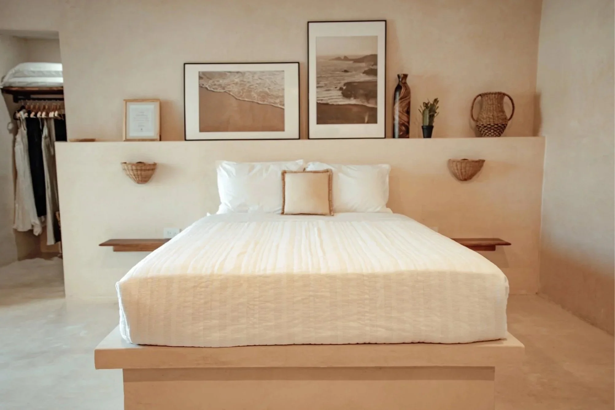 A neatly made bed with white and beige striped bedding, a small beige pillow, and closed white pillows behind it. The headboard area has framed beach photographs and decorative vases, with woven baskets and plants on either side. There is an open clo