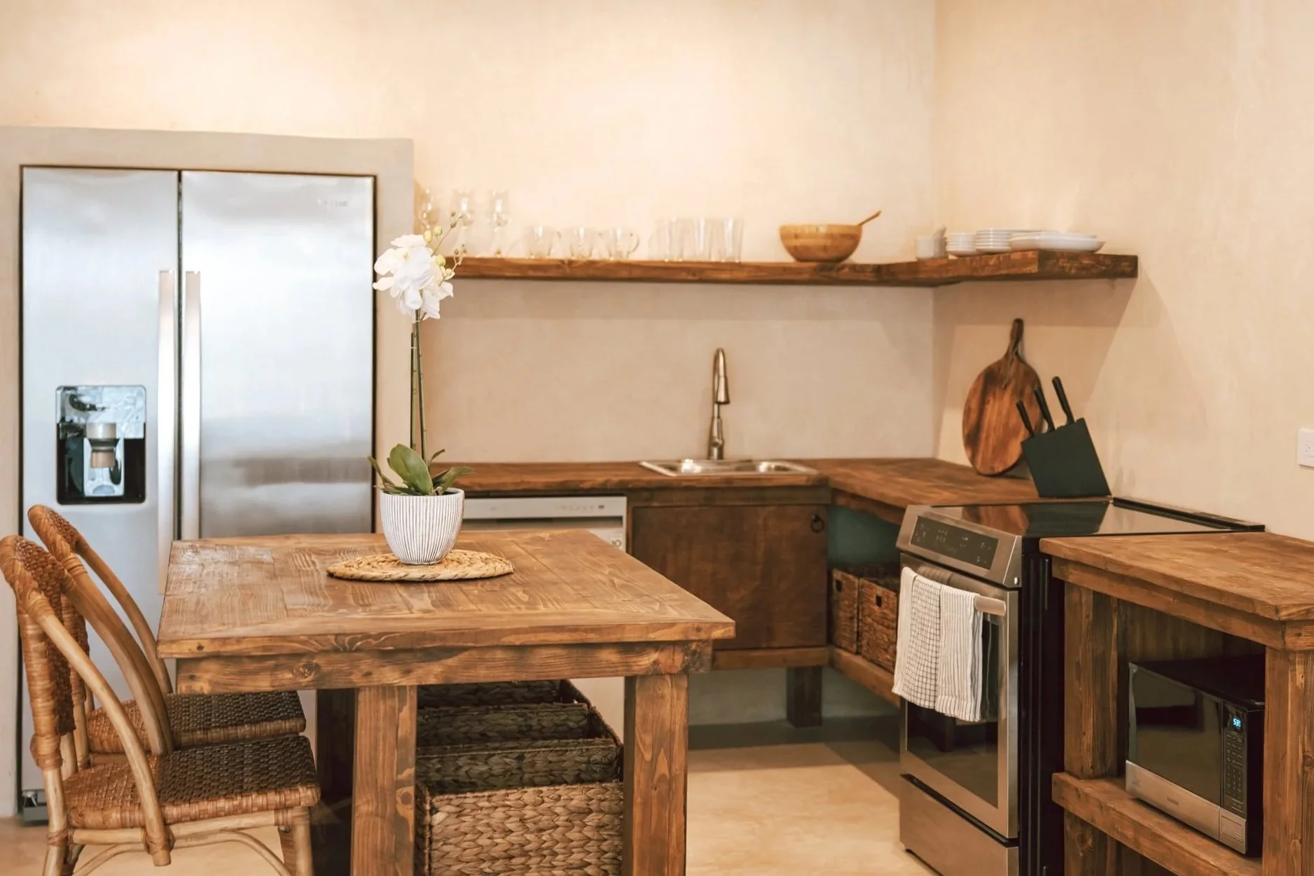 Rustic kitchen with wooden tables, wicker chairs, a stainless steel refrigerator, microwave, and oven, with open shelves holding dishes and glassware, and a potted orchid on the dining table.