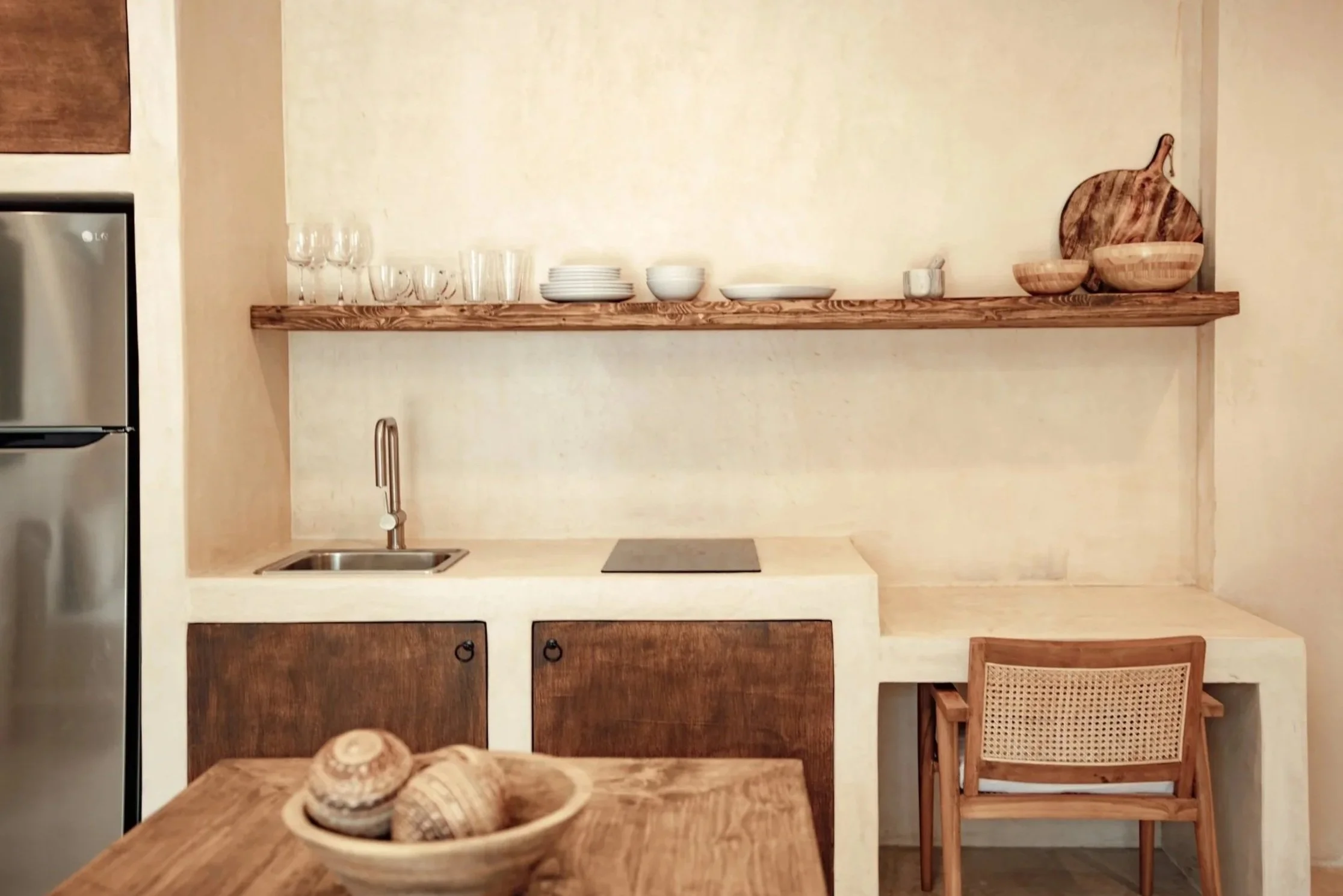 Minimalist kitchen with a small stainless steel sink, a wooden countertop, open shelving with plates and bowls, wooden cabinetry, and a wooden chair with a woven backrest.