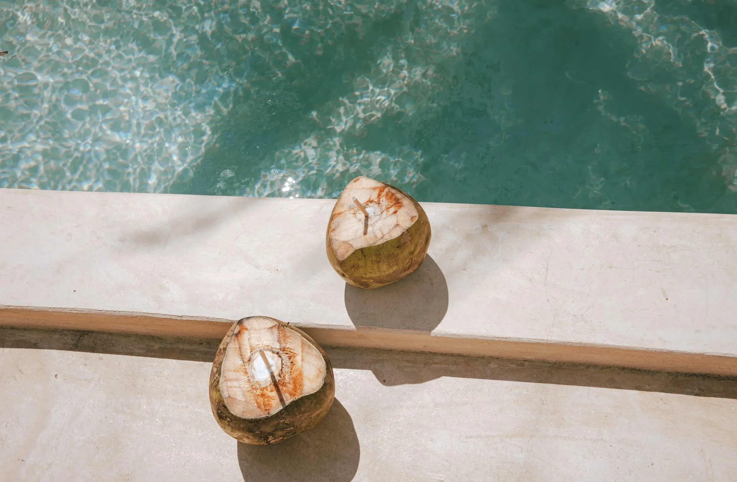 Two coconuts with grated tops placed on a white surface near a swimming pool with clear blue water.