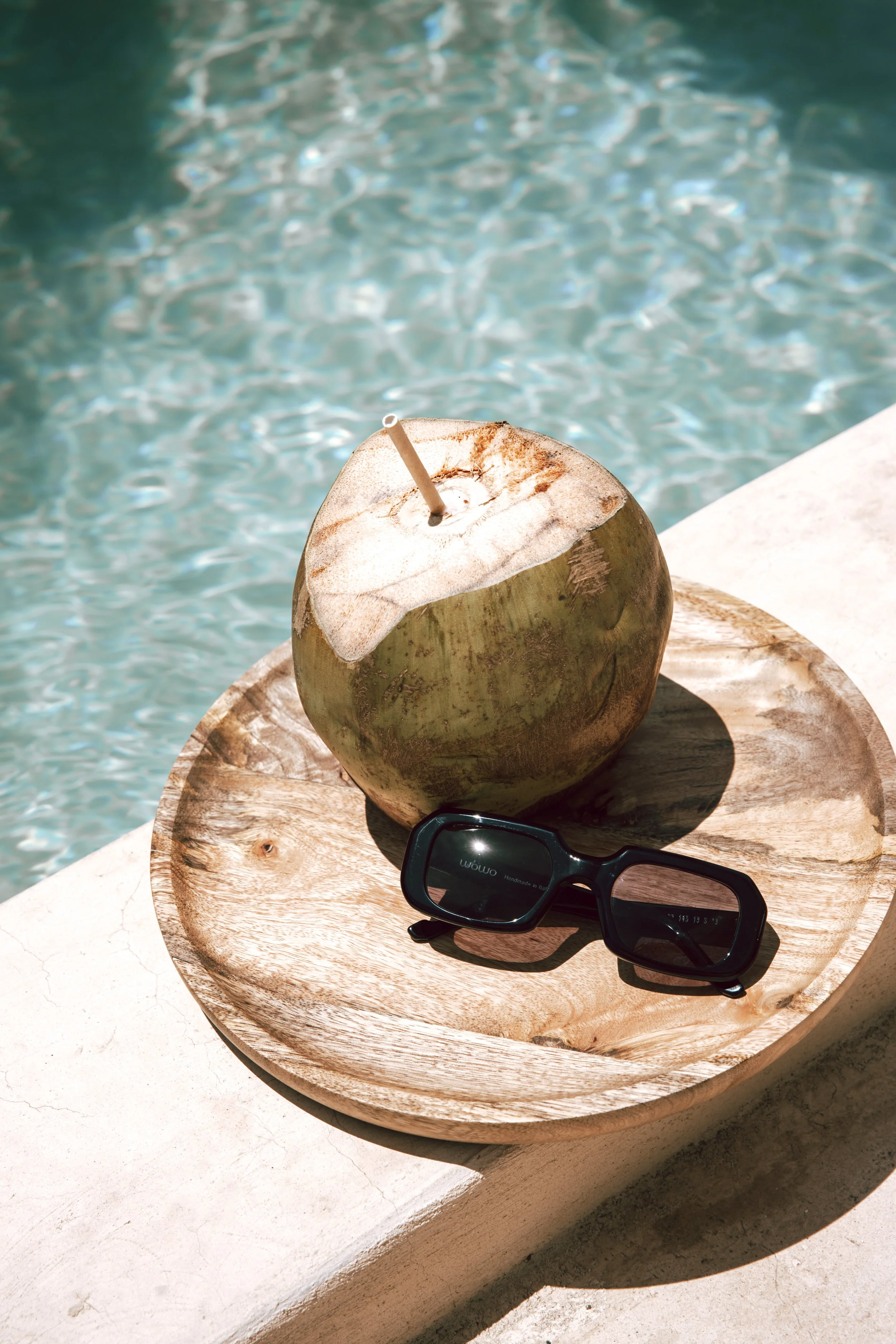A coconut with a straw, black sunglasses, on a wooden tray near a swimming pool.