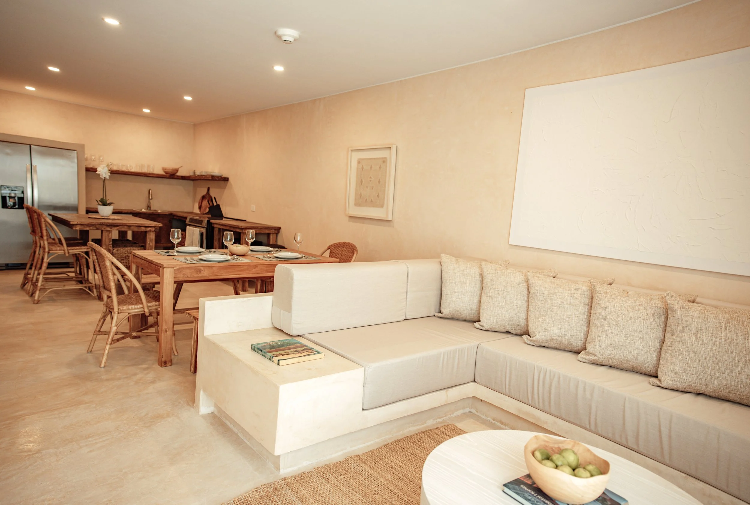 Living and dining area in a modern home with white sectional sofa, beige pillows, wooden dining table set with plates and wine glasses, and a kitchen with a stainless steel refrigerator in the background.