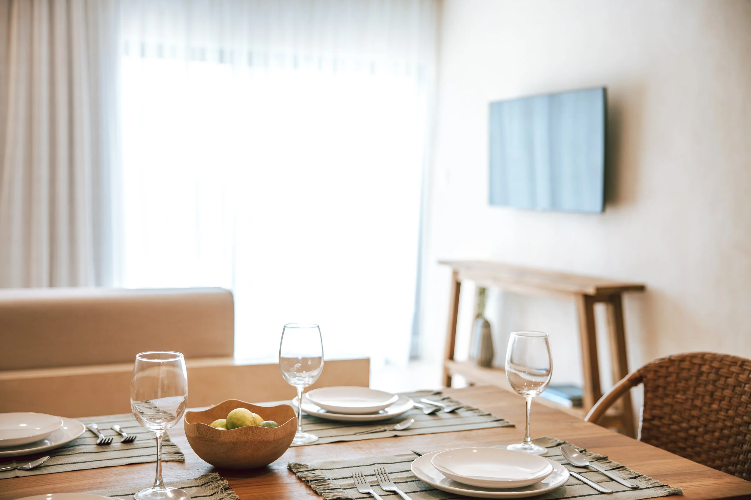 Dining table set with plates, glasses, forks, and a wooden bowl of green apples, in a bright room with window and TV on the wall.
