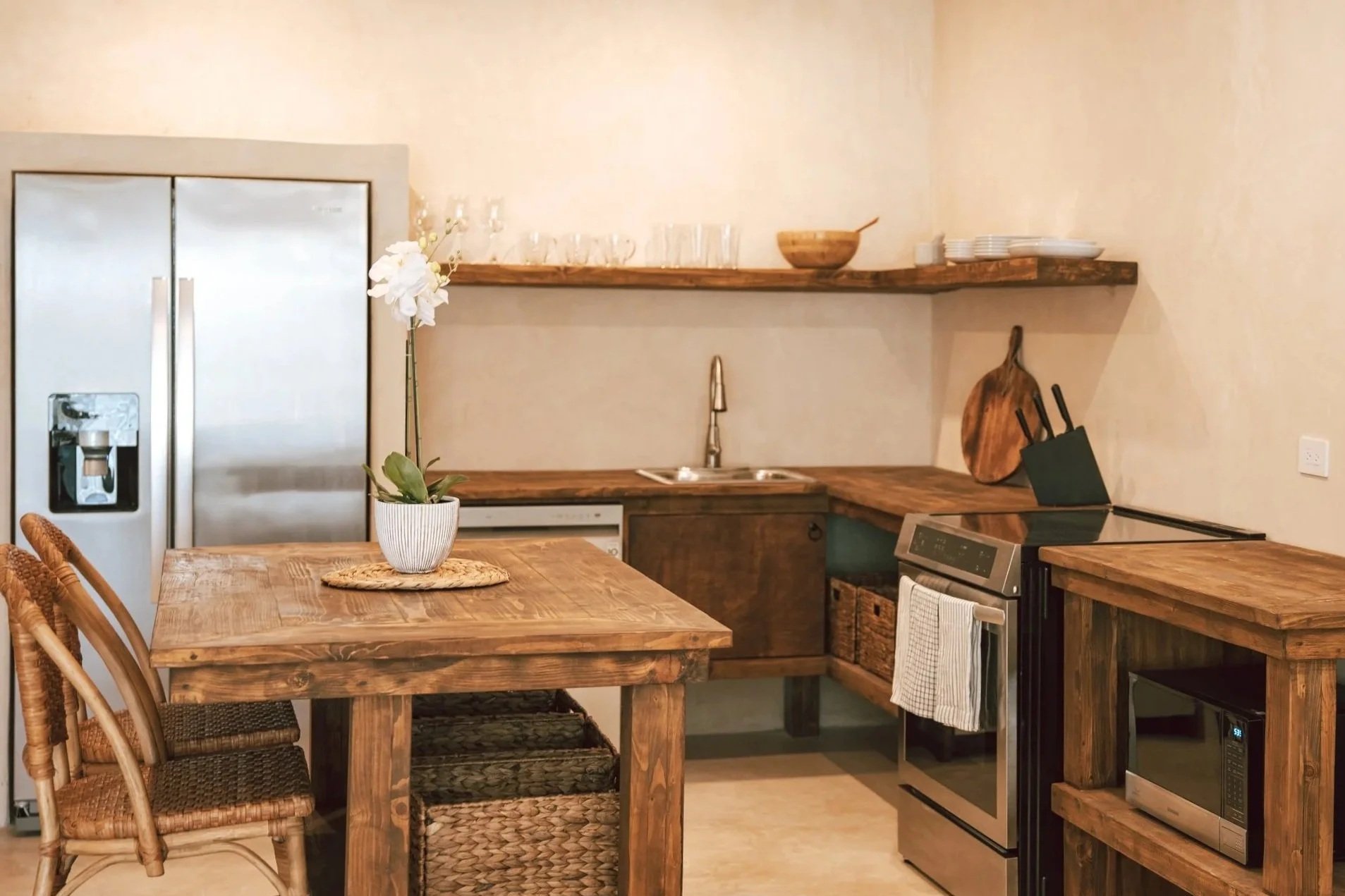 Rustic kitchen with wooden cabinets, a stainless steel refrigerator, a wooden dining table with wicker chairs, a potted orchid, a kitchen counter with a sink, a microwave oven, and open shelves with dishes and bowls.