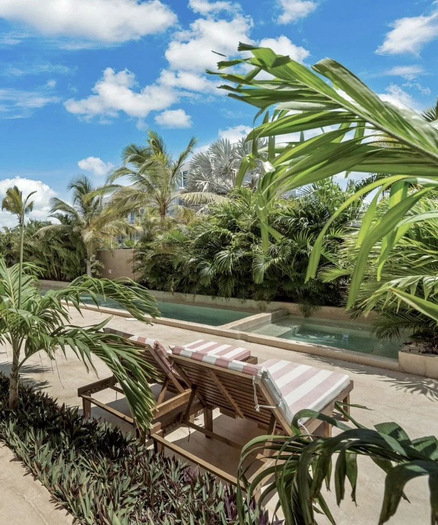 Poolside scene with striped lounge chairs, lush green tropical plants, and a blue sky with white clouds.