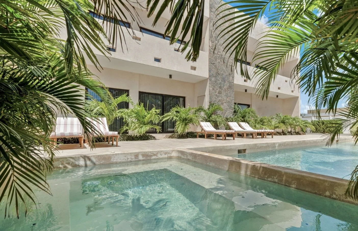 Hotel pool area with lounge chairs, tropical plants, and a building with large windows in the background.