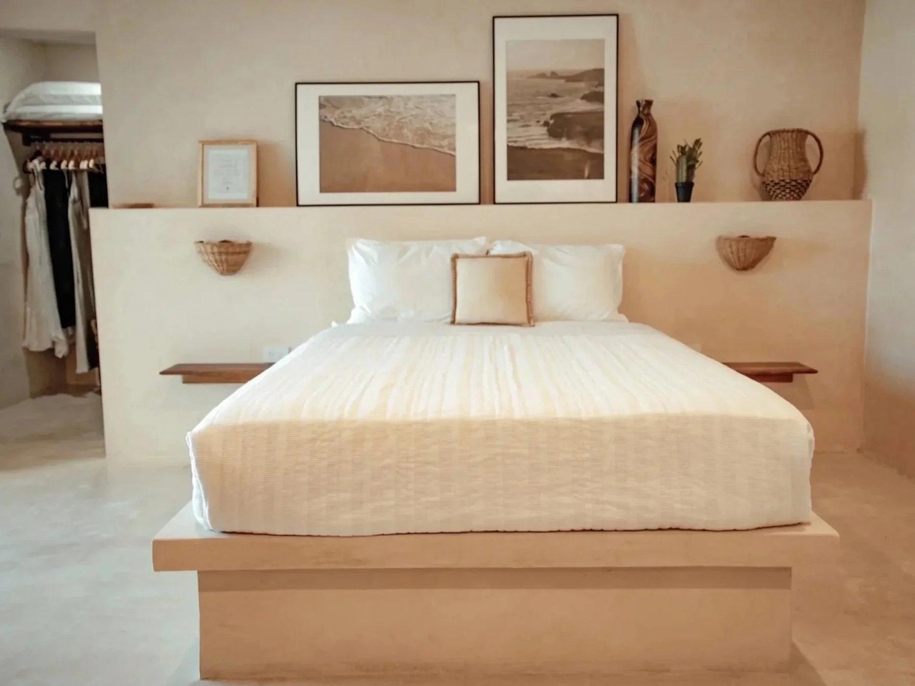 A neatly made bed with white bedding and a small beige pillow, set against a beige wall decorated with framed beach photographs and woven baskets, in a cozy, minimalist bedroom.
