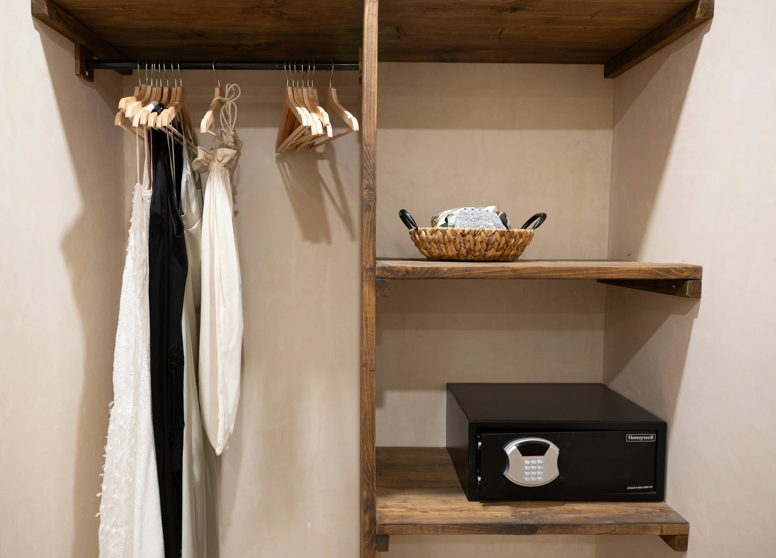 Open closet with a hanging rod holding three dresses or robes, a variety of empty wooden hangers, a woven basket with folded clothes, and a black safe on a wooden shelf.