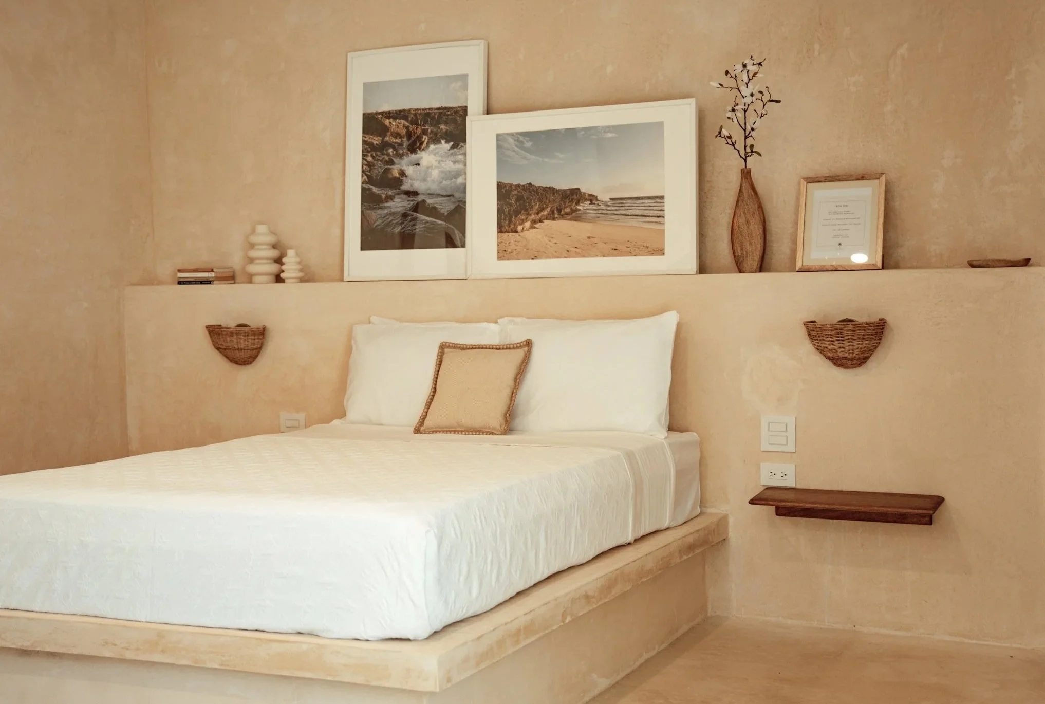 A bedroom with a bed, white bedding, and a decorative pillow. Above the bed, there are framed beach photos, a vase with branches, a certificate, and decorative objects on a shelf. Woven baskets are mounted on the wall on either side of the bed.