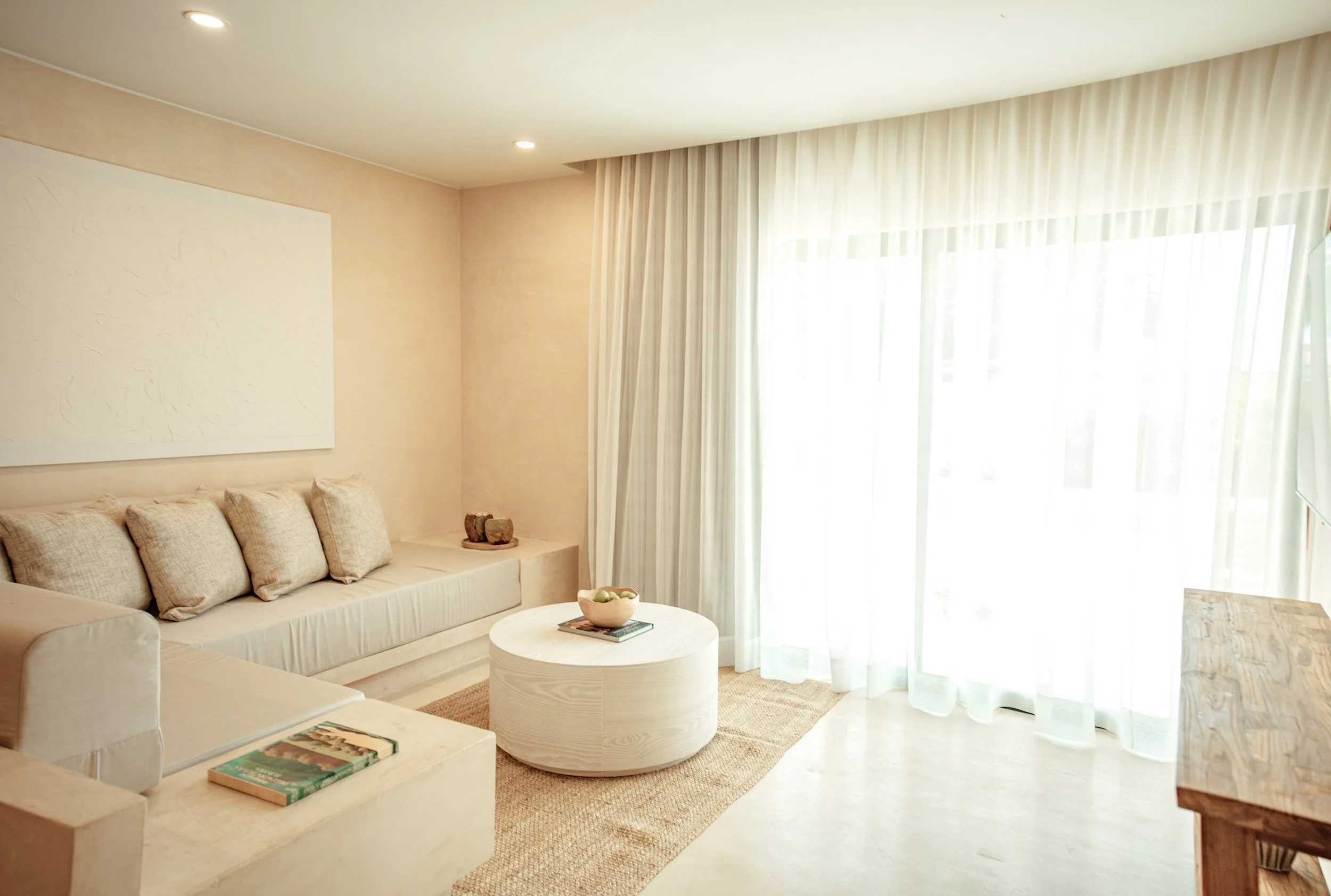 Bright living room with beige walls, cream sofa with matching pillows, white round coffee table, sheer curtains, and sunlight coming through large sliding glass doors.